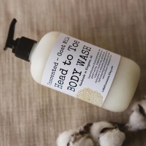 Op de afbeelding: Een witte plastic fles met een zwarte pomp. De fles is gelabeld met "Unscented Goat Milk Head to Toe BODY WASH" en "Handmade in Kingston, MI". Het etiket vermeldt ook de ingredi&euml;nten, waaronder geitenmelk, biologische olijfolie en andere natuurlijke ingredi&euml;nten.
