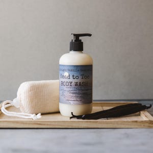 Op de afbeelding: Een doorzichtige fles Midnight Vanilla Goat Milk Head to Toe Body Wash met een zwarte pompdispenser. De fles staat op een houten dienblad met een witte washandje en vanillebonen.
