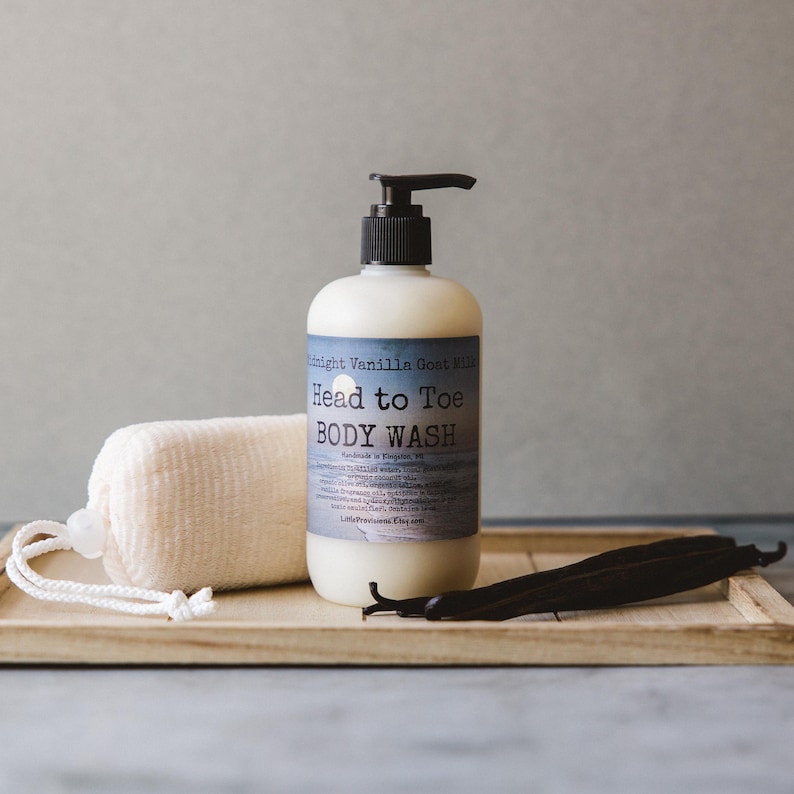 Op de afbeelding: Een doorzichtige fles Midnight Vanilla Goat Milk Head to Toe Body Wash met een zwarte pompdispenser. De fles staat op een houten dienblad met een witte washandje en vanillebonen.