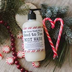 Op de afbeelding: Een witte fles Candy Cane Goat Milk Head to Toe Body Wash met een rood-wit gestreept etiket. De fles is omringd door rode en witte snoepstokken en pepermuntjes.