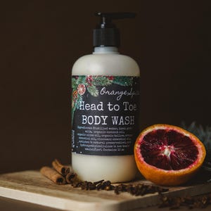Op de afbeelding: Een witte fles douchegel met een zwarte pomp en een etiket met de tekst "Orange Spa Head to Toe Body Wash". De fles staat op een houten oppervlak met een bloedsinaasappel, kaneelstokjes en groenblijvende takken.