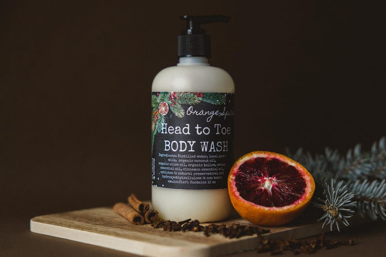 Op de afbeelding: Een witte fles douchegel met een zwarte pomp en een etiket met de tekst "Orange Spa Head to Toe Body Wash". De fles staat op een houten oppervlak met een bloedsinaasappel, kaneelstokjes en groenblijvende takken.