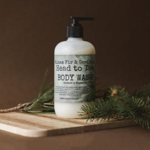 Op de afbeelding: Een doorzichtige plastic fles met Balsam Fir & Goat Milk Head to Toe Body Wash. De fles heeft een zwarte pomp en een wit etiket met groene tekst. De fles staat op een houten oppervlak met een takje groenblijvende.