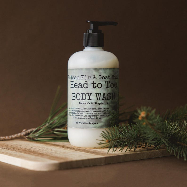 Op de afbeelding: Een doorzichtige plastic fles met Balsam Fir & Goat Milk Head to Toe Body Wash. De fles heeft een zwarte pomp en een wit etiket met groene tekst. De fles staat op een houten oppervlak met een takje groenblijvende.