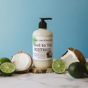 Op de afbeelding: Een doorzichtige plastic fles met kokos-limoen en geitenmelk head-to-toe body wash. De fles heeft een zwarte pomp en een wit etiket met zwarte tekst. De fles staat op een houten schijf met limoenen en kokosnoten eromheen.