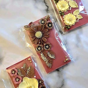 May include: Three rectangular, packaged chocolate bars with floral decorations. The bars are a reddish-brown color and adorned with yellow, brown, and gold-colored flowers and leaves. Each bar is individually wrapped in clear plastic.