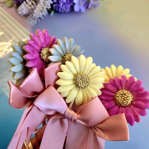 May include: A decorative hair comb with daisy-shaped flowers in pink, yellow, and blue, with gold centers. The comb is decorated with pink satin bows, suitable for weddings or special events. The flowers are in various shades.