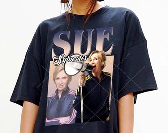 May include: Black t-shirt featuring a graphic print of Sue Sylvester from Glee. The graphic shows a photo of Sue Sylvester holding a megaphone with the text "SUE Sylvester" above the image.