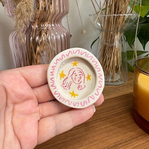 Puede incluir: Un pequeño plato redondo de cerámica con interior crema y borde ondulado rosa. El plato presenta una ilustración de dibujos animados de la cara de una mujer y estrellas amarillas. El plato está sostenido en la mano de una persona.
