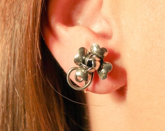 May include: Silver clover-shaped earrings with a small ball in the center. The earrings are worn in a person's ear.