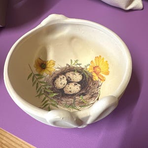 May include: A white, shell-shaped decorative bowl with a painted design. The design features a bird's nest with speckled eggs, yellow flowers, and green foliage. The bowl has a textured surface and a curved edge.