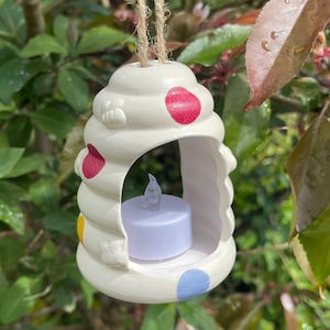 May include: A white ceramic lantern with a swirl design and colourful polka dots in red, yellow, and blue. It contains a battery-operated candle and is suspended by a jute rope.