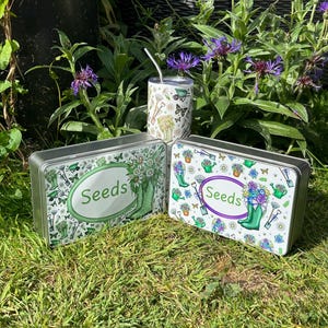 May include: Two rectangular metal seed tins with the word "Seeds" in an oval, and a white tumbler with a straw. The tins and tumbler are decorated with gardening tools, butterflies, and flowers. The tins are on a bed of grass.