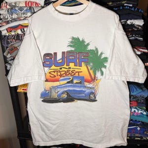 May include: White t-shirt with a graphic print. The design features the words "SURF STREET" in blue and red, a blue pickup truck with a surfboard, palm trees, and a sunset backdrop.