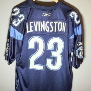 May include: A dark blue sports jersey featuring the name LEVINGSTON and the number 23 in white with light blue outlines on the back. The Reebok logo is visible at the neckline and on the sleeves, which also display a circular logo with the letter A.
