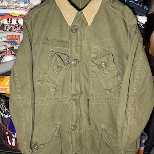 Op de afbeelding: Olijfgroene jas in militaire stijl met een bruin corduroy kraag. De jas heeft meerdere voorzakken, knoopsluitingen en een trekkoord in de taille. De jas hangt aan een houten hanger.