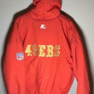 Könnte beinhalten: Eine rote Kapuzenjacke mit dem Schriftzug 49ERS in goldenen Buchstaben auf dem Rücken. Die Jacke hat ein weißes Starter-Logo am Halsausschnitt und am linken Ärmelbündchen sowie einen NFL-Aufnäher am rechten Ärmel und weiße gestreifte Abschlüsse.