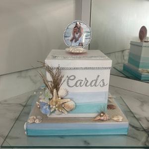 May include: A white box with a blue and white ombre design and the word "Cards" written on it. The box is decorated with seashells and pearls. A photo of a couple is displayed on top of the box.