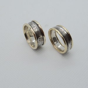 Puede incluir: Dos anillos con un diseño plateado y dorado. Los anillos tienen una banda plateada con un patrón texturizado y un borde dorado. Los anillos están sobre un fondo blanco.