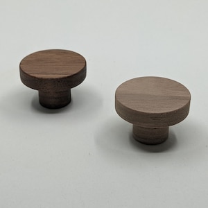 May include: Two wooden cabinet knobs. One is a dark brown color, and the other is a lighter brown. Each knob has a circular top and a short, cylindrical base. The knobs are likely for furniture or decorative use.