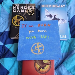 May include: Image of three books and a blue sign with text from The Hunger Games series. The books are "The Hunger Games" and "Mockingjay". The sign reads "If we BURN you burn with US!" and features the series' symbol.