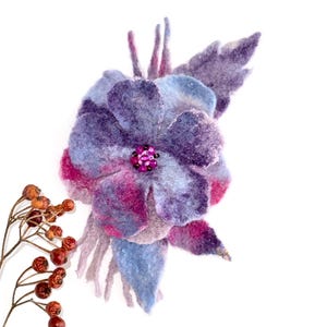 May include: A felted flower brooch featuring layered petals in shades of purple, blue, and magenta. The center is adorned with small dark pink beads. A sprig of dried red berries on a thin brown stem is positioned to the left of the flower.