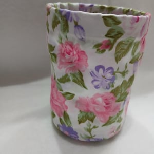 May include: A cylindrical fabric pouch featuring a white background with a repeating floral pattern of pink roses, purple blossoms, and green leaves. The top edge is gathered with a simple hem, creating a soft, rounded opening for the container.
