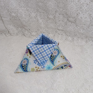 May include: A triangular fabric basket with a blue and white gingham interior. The exterior features a white paisley pattern with blue, yellow, and green floral accents. The basket is open at the top.