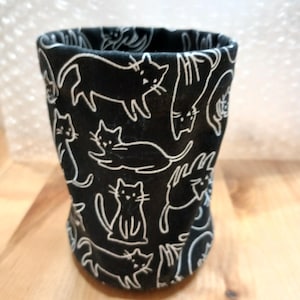 May include: A black fabric cup with a white cat pattern. The cats are in various poses, outlined in white against the black background. The cup is cylindrical with a slightly flared top, and is sitting on a wooden surface.