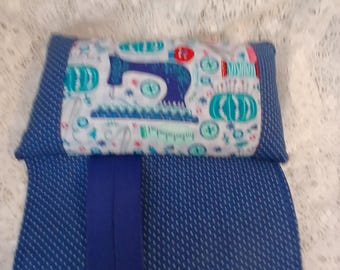 May include: A blue sewing machine themed fabric case with a blue and white dotted pattern. The case has a flap with a sewing machine design, spools, and buttons. A solid blue band runs down the center.