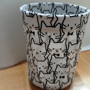 May include: A cylindrical fabric container featuring a repeating pattern of white cats with black outlines. Each cat is depicted with whiskers, pointed ears, and a small black bow tie. The fabric has a soft, flexible structure with a circular base.