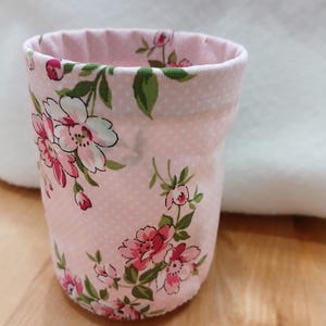 May include: A small, fabric-covered container with a pink floral pattern. The container is cylindrical with a slightly flared top. The pink fabric has a white polka dot pattern and is decorated with pink and white flowers and green leaves.