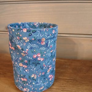 May include: A cylindrical fabric container featuring a blue background with a repeating pattern of small pink flowers, green leaves, and thin stems. The fabric is folded over at the top rim, revealing the interior lining with the same floral print.