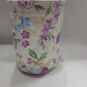 May include: A cylindrical fabric container featuring a cream-colored background with a repeating floral pattern. The design includes small purple, light blue, and orange flowers with green leaves and stems scattered across the entire exterior surface.