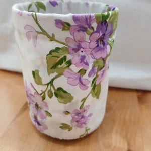 May include: A fabric-covered cylindrical container with a floral pattern. The white fabric is adorned with purple flowers, green leaves, and stems. The container is likely a pencil holder or a small storage item.