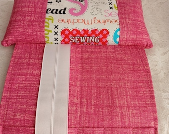 May include: A pink sewing machine needle holder with a white ribbon and a decorative fabric band. The band features the word "SEWING" in red and white, along with other sewing-related text and graphics.