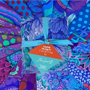 Può includere: Una collezione di quadrati di tessuto fantasia nei toni del blu, viola e verde acqua. Il tessuto è legato con un nastro azzurro e presenta un'etichetta con la scritta "KAFFE FASSETT COLLECTIVE for Free Spirit". I tessuti hanno disegni floreali e astratti.