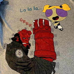 May include: A gray t-shirt featuring cartoon characters Hellboy and Buttercup. Hellboy is red with a black coat and the text "Grrr". Buttercup is yellow, purple, and green. The text "La la la..." is above the characters.