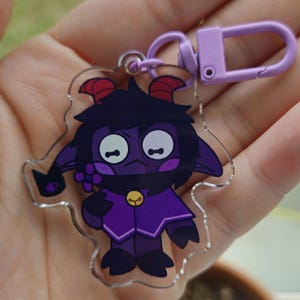 May include: A clear acrylic keychain featuring a cartoon character with purple skin, black hair, and red horns. The character wears a purple outfit and holds a flower. The keychain has a purple clasp.