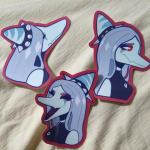 Puede incluir: Tres pegatinas con un personaje estilizado con una nariz larga, cabello gris y un sombrero azul y blanco. El personaje tiene maquillaje rojo y azul y está delineado en rojo. Las pegatinas están sobre una superficie beige.