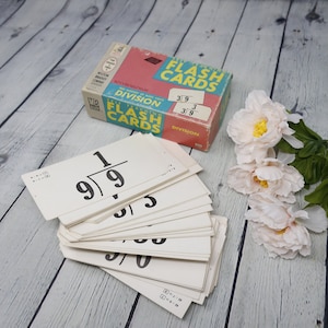 Puede incluir: Un juego de tarjetas de memoria vintage para aprender las tablas de división. Las tarjetas son blancas con números negros y una caja azul y roja con el texto "Self Teaching Flash Cards Division 3/9 3 3/9".