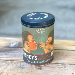 May include: A vintage tin container with a blue lid and a colorful illustration of a couple sharing a Hershey's chocolate kiss. The text on the tin reads "A Kiss for You" and "Hershey's Milk Chocolate".