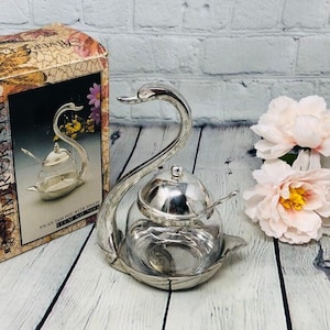 Könnte beinhalten: Silberner Schwan Zuckerdose und Löffelset mit einer klaren Glasschale. Das Set ist in einer Schachtel mit einem Blumendesign verpackt und die Worte "Silver Crest" sind auf der Seite aufgedruckt.