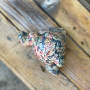 May include: A floral patterned ceramic bear figurine with a dark blue background and pink, white, and yellow flowers. The bear is sitting on a wooden surface.