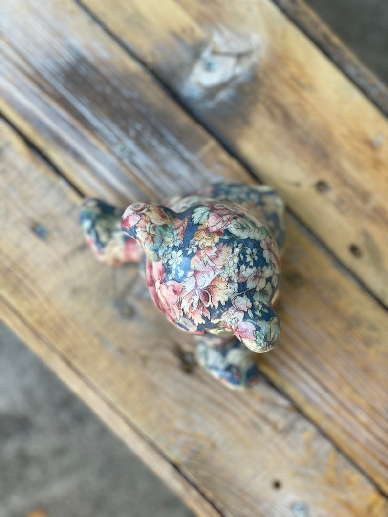 May include: A floral patterned ceramic bear figurine with a dark blue background and pink, white, and yellow flowers. The bear is sitting on a wooden surface.