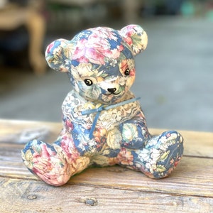 May include: A ceramic teddy bear figurine covered in a floral fabric with a blue ribbon around its neck. The bear is sitting on a wooden surface.