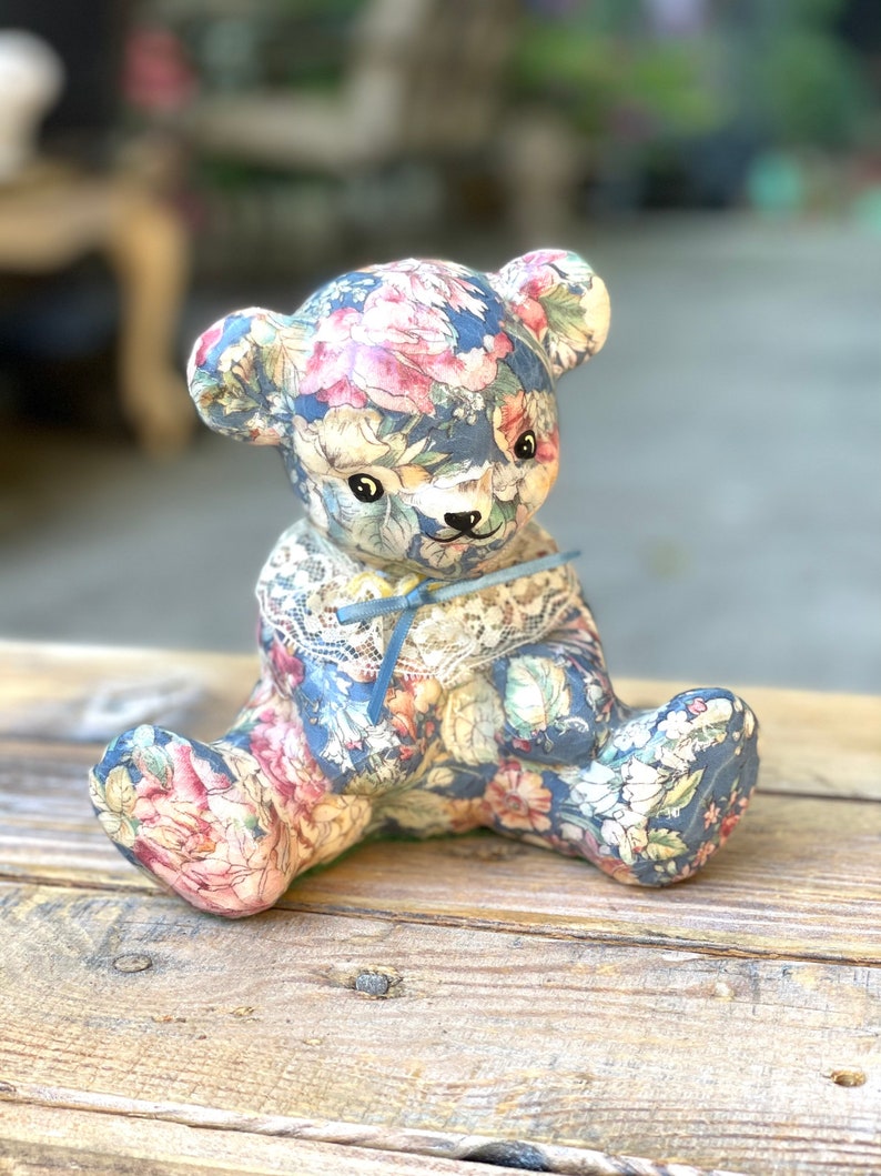 May include: A ceramic teddy bear figurine covered in a floral fabric with a blue ribbon around its neck. The bear is sitting on a wooden surface.