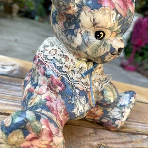 May include: A ceramic teddy bear figurine with a floral pattern in shades of blue, pink, and white. The bear is wearing a lace collar with a blue ribbon.