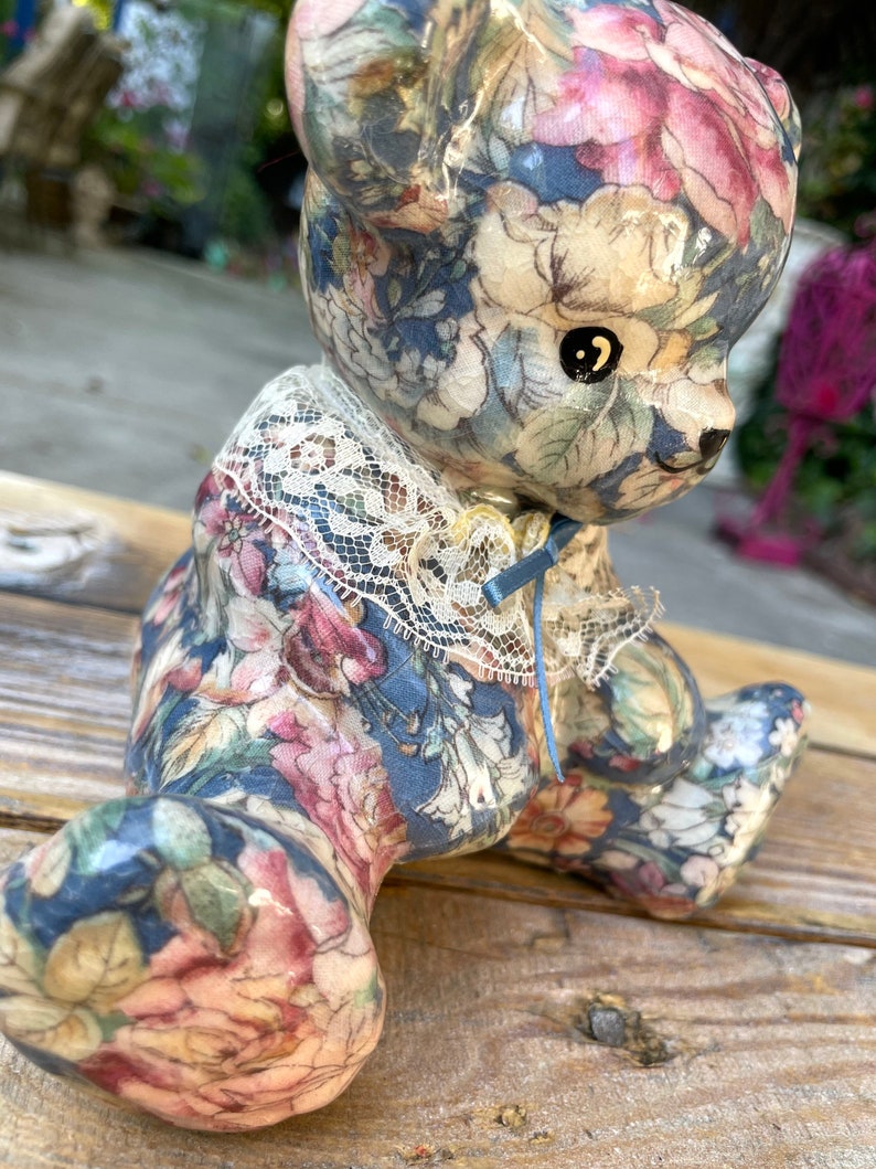 May include: A ceramic teddy bear figurine with a floral pattern in shades of blue, pink, and white. The bear is wearing a lace collar with a blue ribbon.