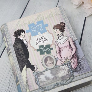 May include: A vintage-style jigsaw puzzle box featuring a couple in period clothing. The box is decorated with a floral pattern and the text "Jigsaw Puzzle Jane Austen 500 Pieces".
